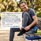 A man sitting on a wooden park bench wearing a dark gray t-shirt and gray running shoes, with one foot in a black sock while the other shoe lies beside him. A blue foot support pad is placed on the bench next to the shoe.