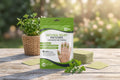 A stand-up pouch of natural relief patches for hands and fingers sits on a wooden table outdoors, with a stack of square patches, a small plant in a basket, and a sprig of herbs against a blurred garden background.