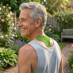 A smiling older man in a gray tank top stands in a garden, showing a green neck and shoulder support pad applied to the back of his neck.